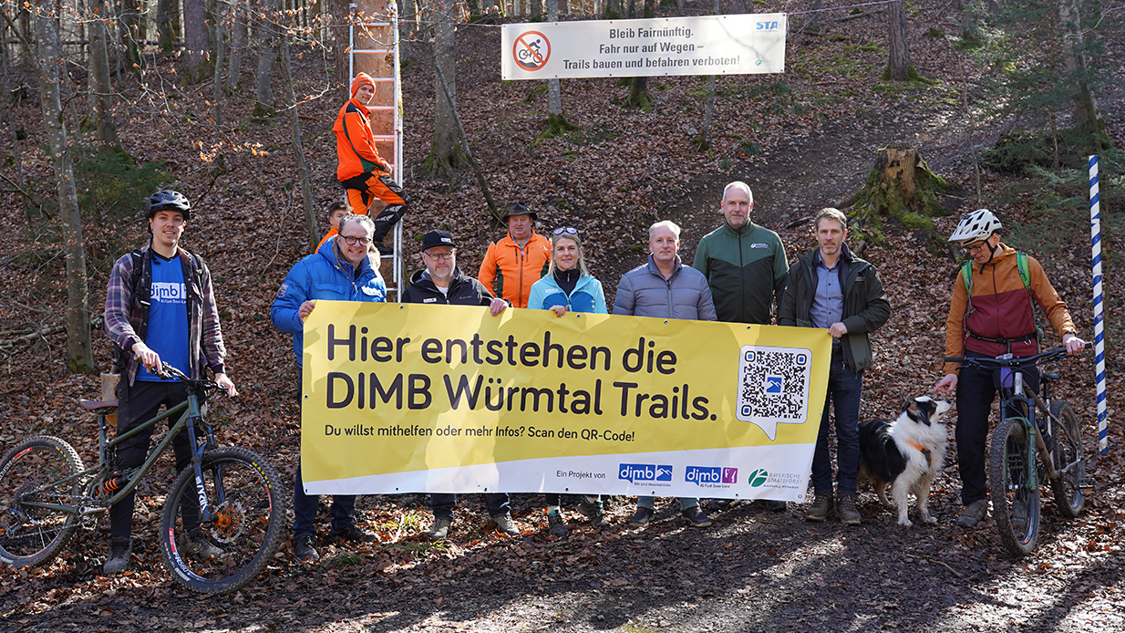 Mitarbeiter des Forstbetriebs und Mountainbiker stehen mit einem Banner, auf dem für die Würmtal-Trails geworben wird, im Wald